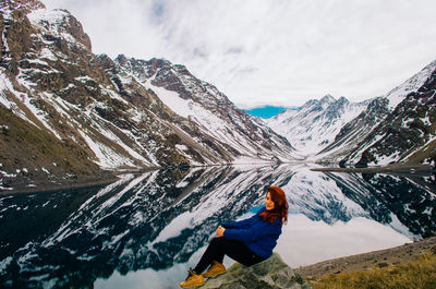 Full length of woman sitting by lake against snowcapped mountain