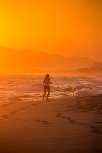 Silhouette woman running on beach against orange sky during sunset