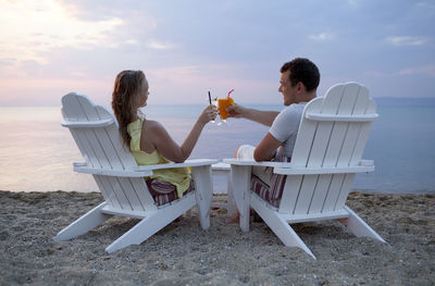 Rear view of couple toasting drinks while sitting on beach