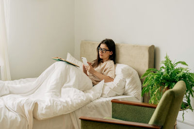 Female pretty student reading a book and taking notes on mobile phone while lying in bed