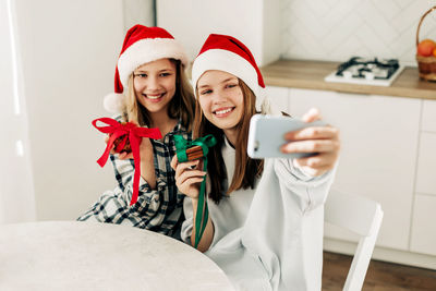 Portrait of two cute sisters in a santa claus christmas hat in the kitchen, taking photos 