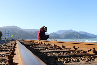 Side view of woman sitting by railroad track against clear blue sky