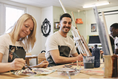 Portrait of happy young man sitting by female student painting at table in art class