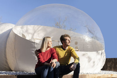 Young man with girlfriend sitting in front of transparent dome hotel on sunny day