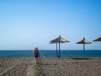 Rear view of woman walking on sandy beach against clear blue sky