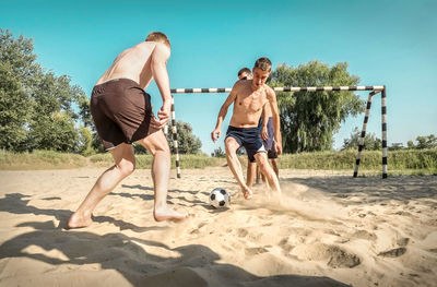 Happy young people in active dynamic action with ball, play in football on sand, summer, sunny day.