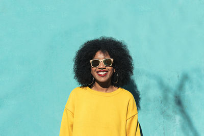 Portrait of smiling young woman wearing sunglasses against standing yellow wall
