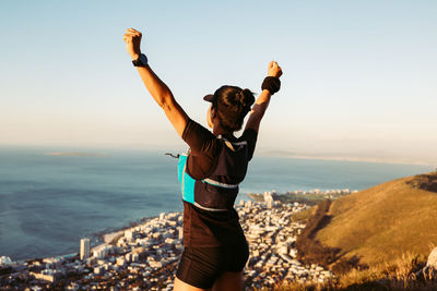 Rear view of woman with arms raised standing on rock against sky during sunset