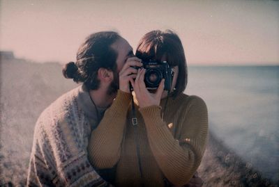 Young man photographing woman standing by sea against clear sky