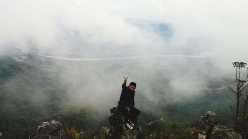 High angle view of man gesturing peace sign while sitting on rock at cliff against mountains during foggy weather