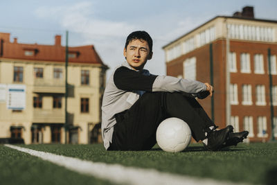 Ambitious teenage boy with soccer ball sitting on sports field