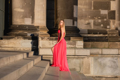 Full length side view portrait of young woman in pink evening gown standing at on steps
