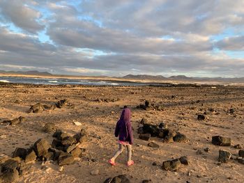 Rear view of girl walking at beach against cloudy sky