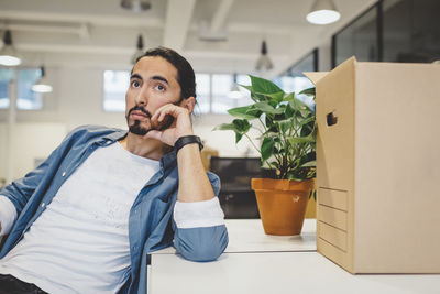 Thoughtful young businessman sitting by cardboard box and potted plant in new office