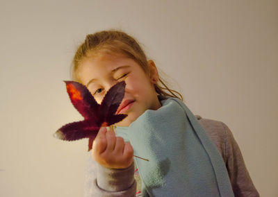Portrait of girl winking while holding maple leaf against wall