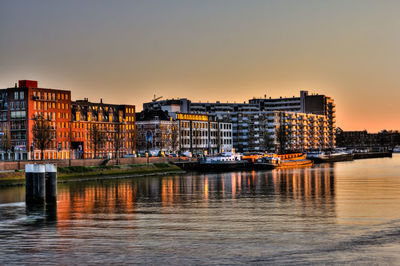 River by illuminated buildings against sky during sunset