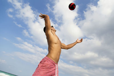 Shirtless man playing volleyball at beach against sky