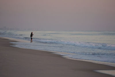 Man fly-fishing while standing on shore at beach during sunset
