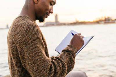 Side view of cropped content african american male taking notes in notepad while sitting on rocky coast near sea in city