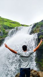 Rear view of young man standing with arms outstretched against waterfall