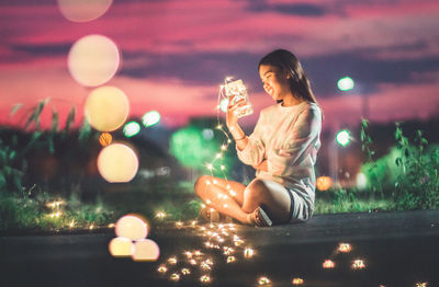 Young man holding jar with illuminated christmas lights while sitting on footpath against sky at night