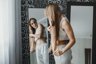 Young woman brushing hair standing against mirror at home