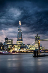 Illuminated tower bridge over river against cloudy sky