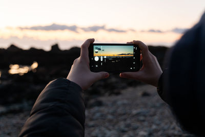 Detail of hands holding smart phone taking photo of sunset over beach