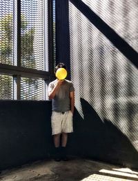 Front view of man holding yellow balloon against windows and wall.