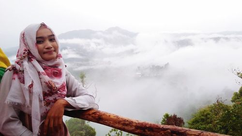 Portrait of young woman in hijab standing by wooden railing on mountain during foggy weather