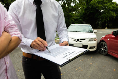 Midsection of insurance agent discussing over agreement with client while standing by cars