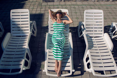 Woman lies on a chaise longue in a green striped dress and a hat by the pool