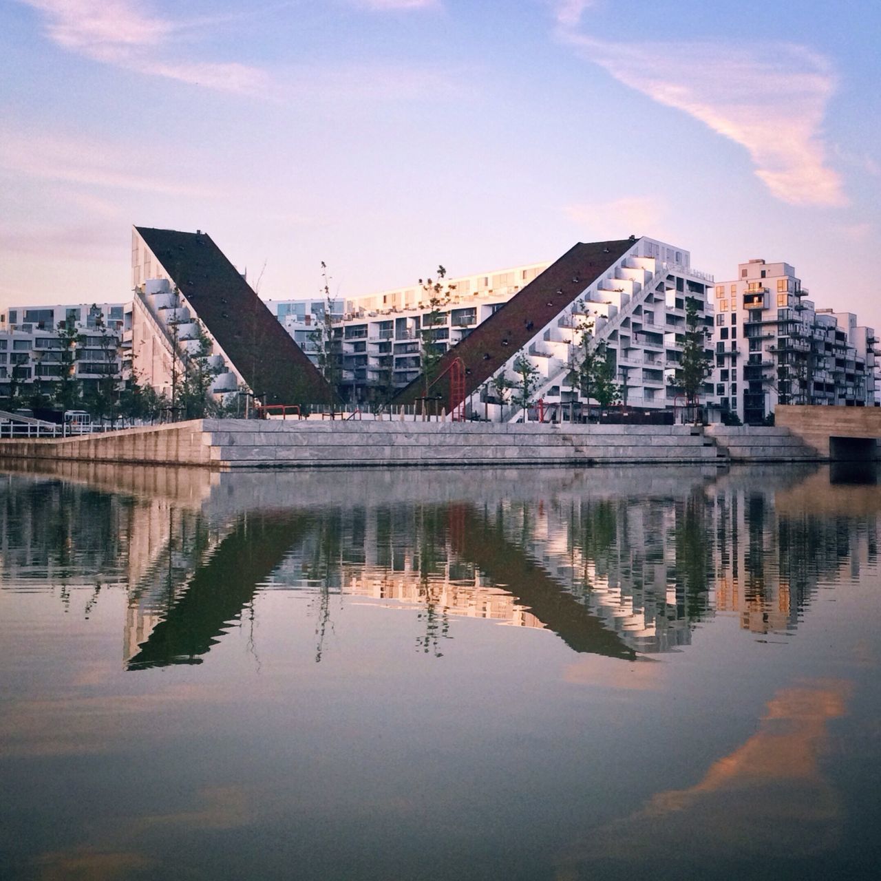 architecture, built structure, reflection, building exterior, water, waterfront, sky, city, river, cloud - sky, lake, standing water, cloud, sunset, outdoors, no people, building, skyscraper, symmetry, modern