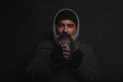 Homeless middle-aged man with a gray beard holding a cup of hot tea to warm himself on a cold night