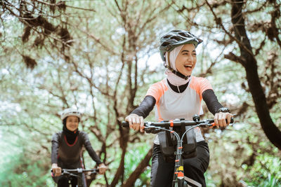 Smiling females riding bicycle against trees