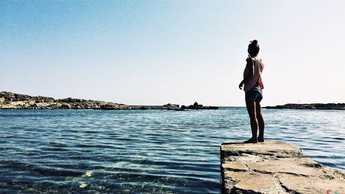 Rear view of woman standing on rock in sea against clear sky