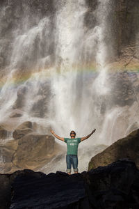 Young man with arms outstretched standing against waterfall