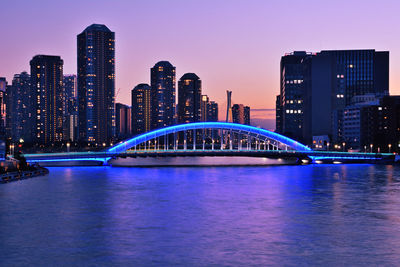 Illuminated bridge over river by buildings in city at dusk