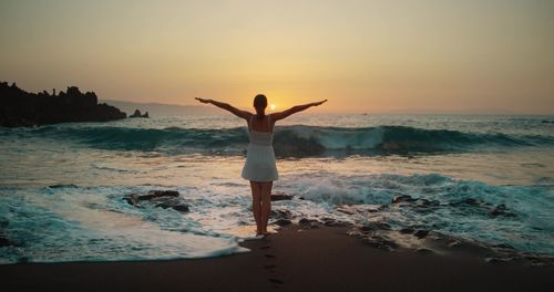 Woman silhouette raises hands up on black sand beach at sunset. twilight orange sky.