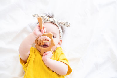 Healthy asian baby girl in yellow bodysuit plays with wooden toy teether for teeth on white bedding