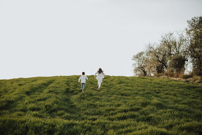 Brother and sister running on meadow against clear sky