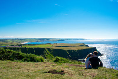 Couple sitting on cliff with sea in background against blue sky