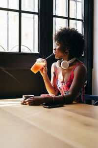 Side view of stylish african american young female with headphones on neck having breakfast and drinking fruit juice while sitting at table next to window and enjoying morning in cafe