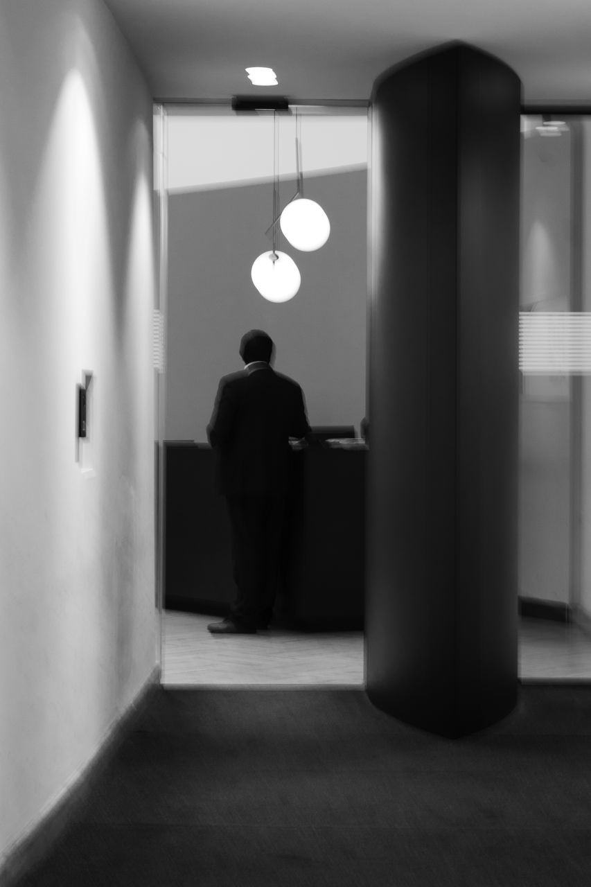 REAR VIEW OF MAN WALKING IN ILLUMINATED CORRIDOR