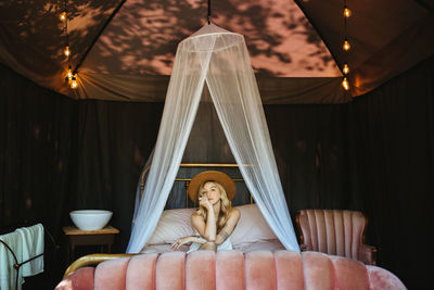 Serene female with stylish hat sitting on comfortable bed with translucent fabric placed on terrace of patio