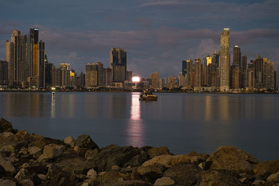 Illuminated buildings by sea against sky