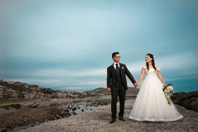 Wedding couple holding hands while standing against cloudy sky
