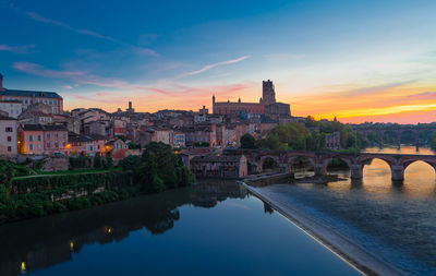 Bridge over river by buildings against sky during sunset