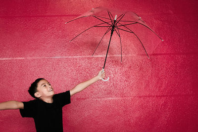 Boy holding umbrella while standing against red wall