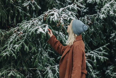 Woman wearing warm clothing looking at fir tree with snow in forest